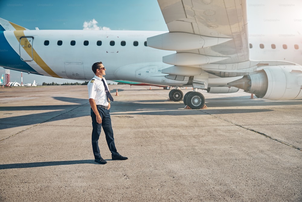 Jovem alegre de uniforme está relaxando ao sol na pista perto do avião de passageiros antes de embarcar