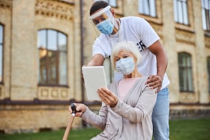 Neugierige ältere Frau in medizinischer Maske sitzt draußen mit einem Tablet und einem jungen Mann, der darauf zeigt