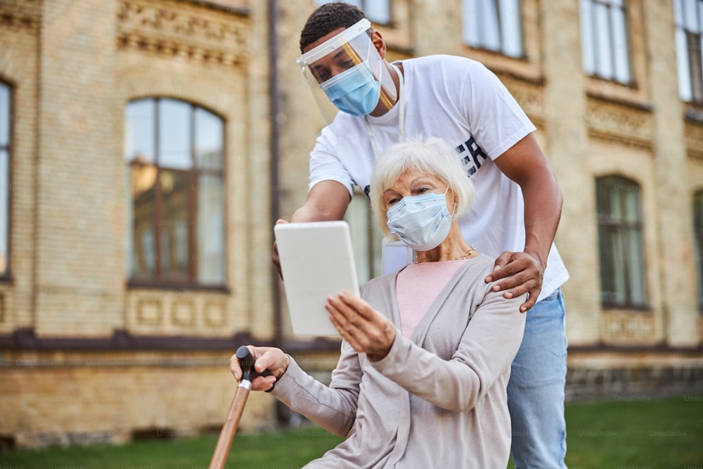 Curious aged woman in medical mask sitting outdoors with a tablet and a young man pointing to it