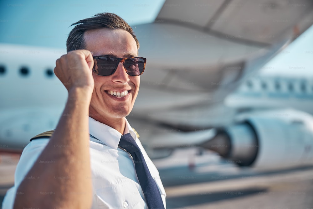 Retrato de un joven profesional masculino guapo sonriente de pie cerca del avión de pasajeros después de la llegada al aeropuerto