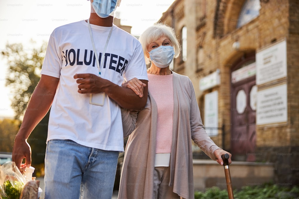 Ruhige ältere Dame mit medizinischer Maske, die einen Gehstock hält, während ein verantwortlicher Freiwilliger Einkäufe in Papiertüten trägt