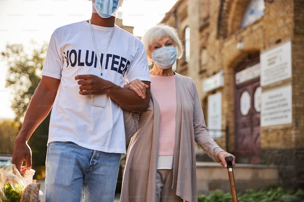 Calm elderly lady wearing a medical mask holding a walking stick while a responsible volunteer carrying purchases in paper bag