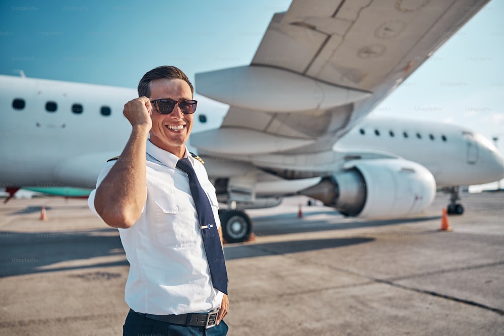 Porträt eines fröhlichen, gutaussehenden jungen Mannes in Uniform, der Freizeit auf dem Laufsteg in der Nähe des Flugzeugs genießt