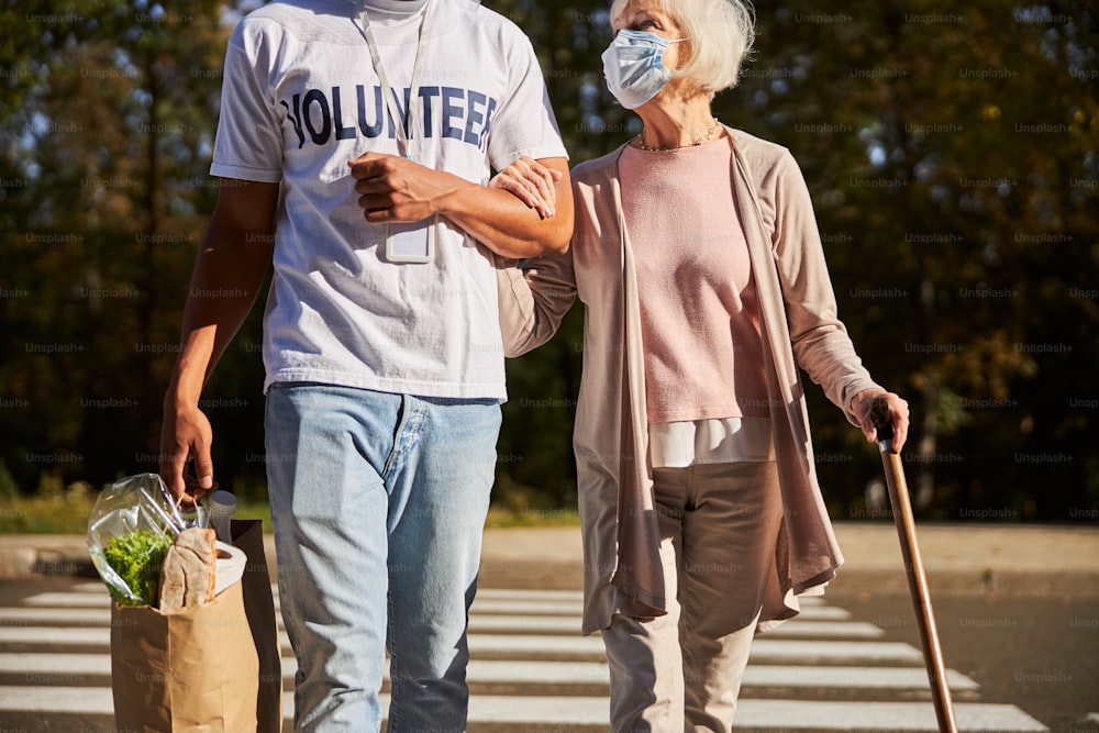 Elderly woman in medical mask looking at the young man by her side carrying shopping bag