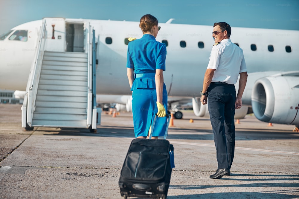 Glücklicher, gutaussehender Pilot und hübsche Stewardess steigen vor dem Flug im Passagierjet mit Gepäck ein