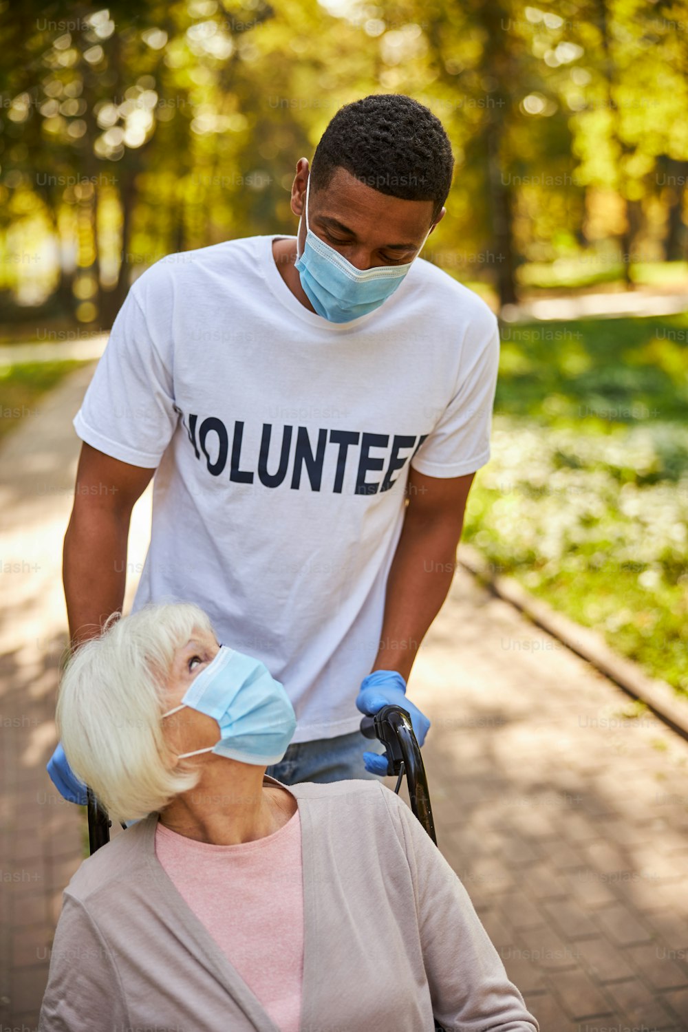 Caring volunteer in a white T-shirt and blue gloves assisting a retired woman and rolling her wheelchair on the road