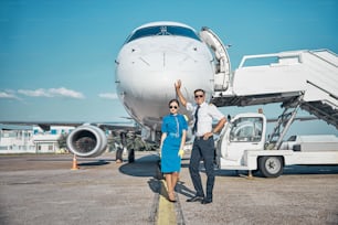 Full length portrait of joyful young pilot and stewardess in sunglasses standing on runway after arrival