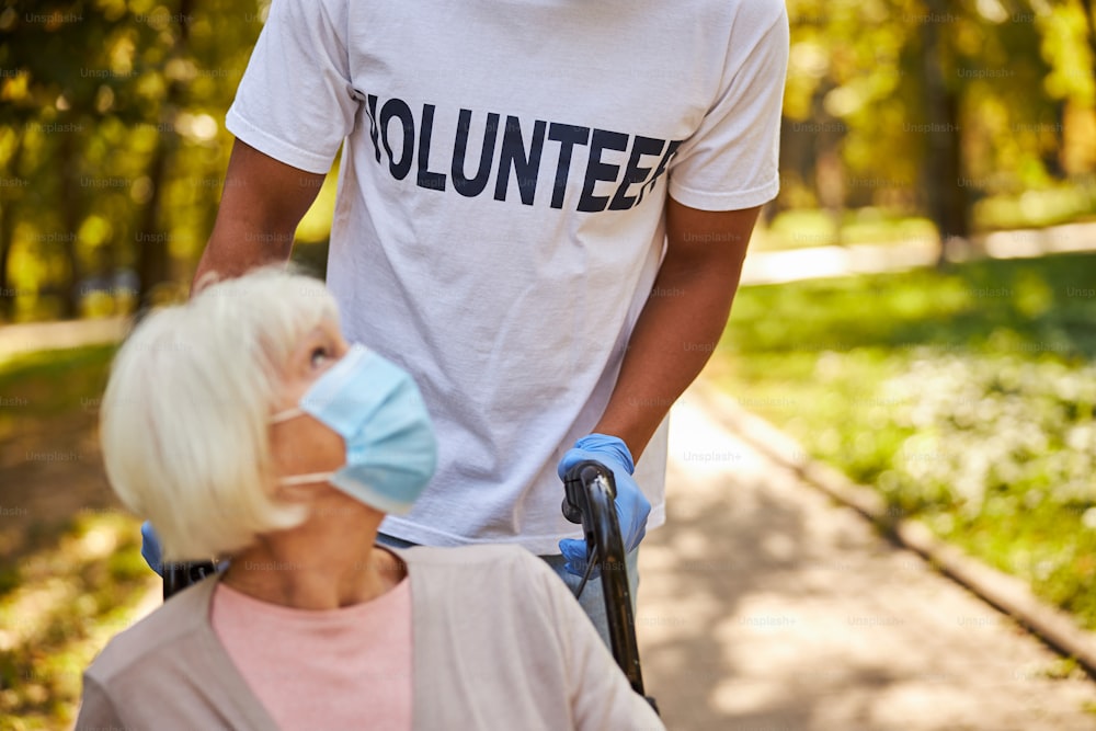 Foto recortada de um voluntário afro-americano usando luvas médicas e segurando as alças de uma cadeira de rodas com uma mulher idosa