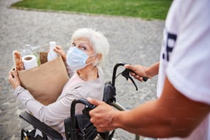 Aged woman with disability wearing a medical mask and holing a paper bag full of purchases while looking at someone pushing her wheelchair