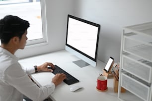 Young businessman typing on keyboard and using computer pc and smart phone with blank screen in home office.