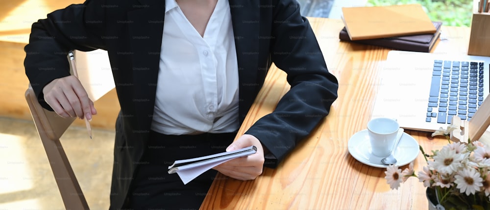 Foto cortada de uma jovem empresária segurando caderno enquanto estava sentada no espaço de trabalho de madeira.