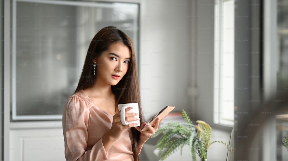 Porträt Asia-Frau mit Kaffeetasse und Smartphone mit Blick in die Kamera.