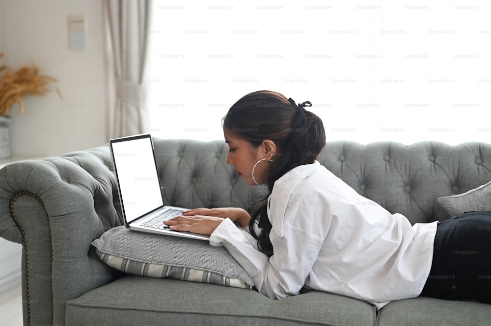 Mujer joven acostada en el sofá mientras lee información en la computadora portátil en casa.