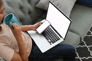 Cropped shot of female freelancer using laptop and smart phone with blank screen on sofa at home.