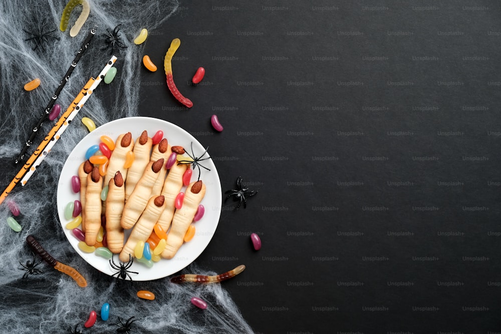 Concept de pâtisserie d’halloween effrayant. Biscuits cuits au four en forme de doigts de main de sorcières, bonbons colorés, toile d’araignée sur fond noir. Pose à plat, vue de dessus