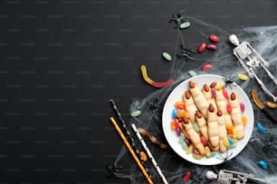 Halloween composition. Flat lay cookies fingers, spiderweb, colorful candies, skeleton on black background. Halloween party concept.