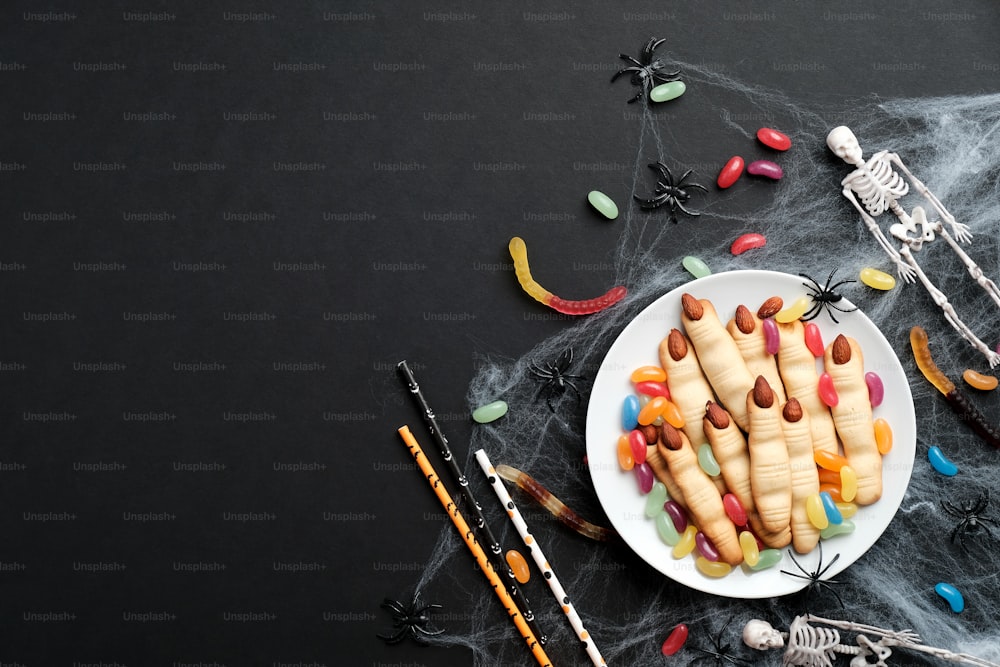 Halloween-Komposition. Flache Kekse Finger, Spinnennetz, bunte Bonbons, Skelett auf schwarzem Hintergrund. Halloween Party-Konzept.