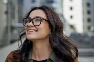 Close up portrait of a young happy charming woman wearing eyeglasses looking aside and smiling while walking city streets, standing against blurred urban background. People lifestyle concept