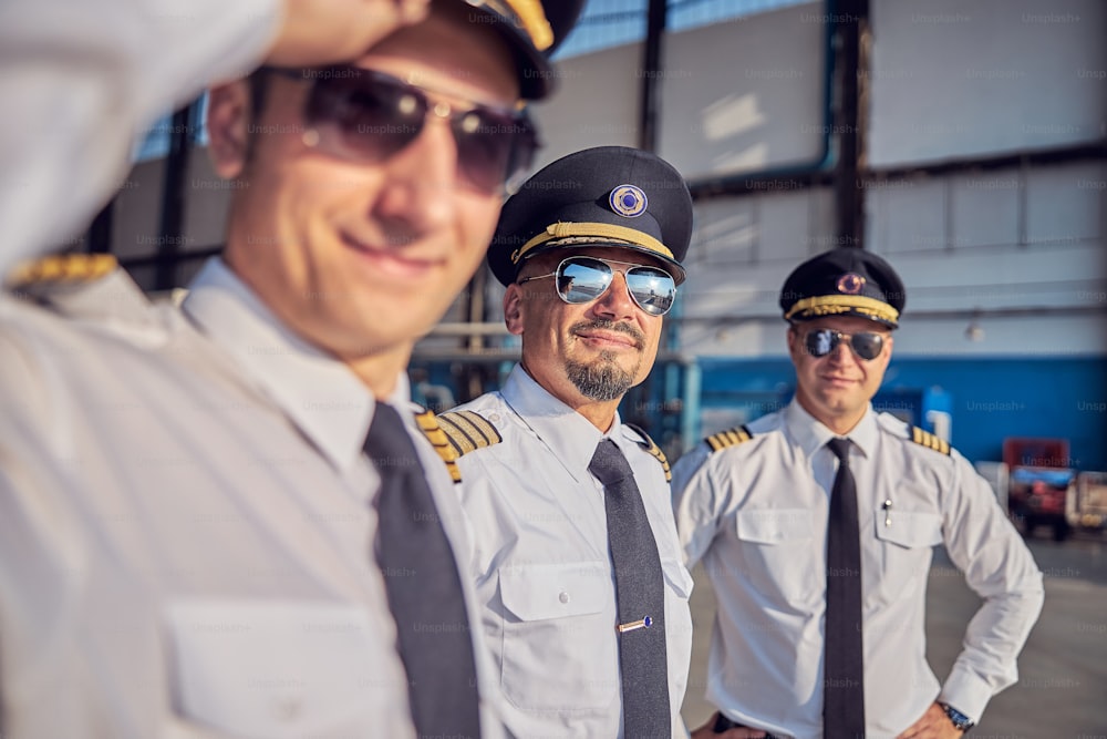 Retrato de cerca de pilotos guapos y seguros de sí mismos con camisa blanca y gafas de sol posando y mirando a la cámara de fotos mientras están de pie en el hangar del aeropuerto
