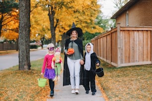 Trick or treat. Mother with children going to trick or treat on Halloween holiday. Mom with kids boy and girl in party costumes with baskets going to neighbour houses for candies and treats.