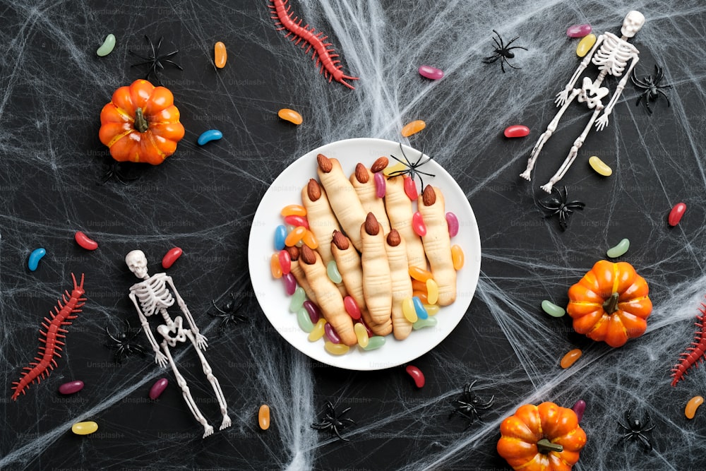Concepto de pastelería de Halloween. Sabrosos dedos de bruja con uñas de almendras, telas de araña, caramelos de colores, esqueletos sobre fondo oscuro. Plano, vista superior.