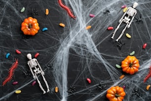 Black halloween background with spider web, colorful candies, pumpkins, skeletons. Flat lay, top view. Happy halloween concept.