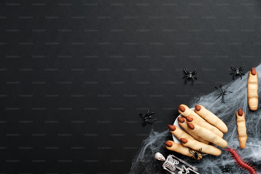 Halloween composition. Witch hand fingers, spiderweb, spiders, skeletons on black background. Flat lay, top view. Halloween pastry concept.