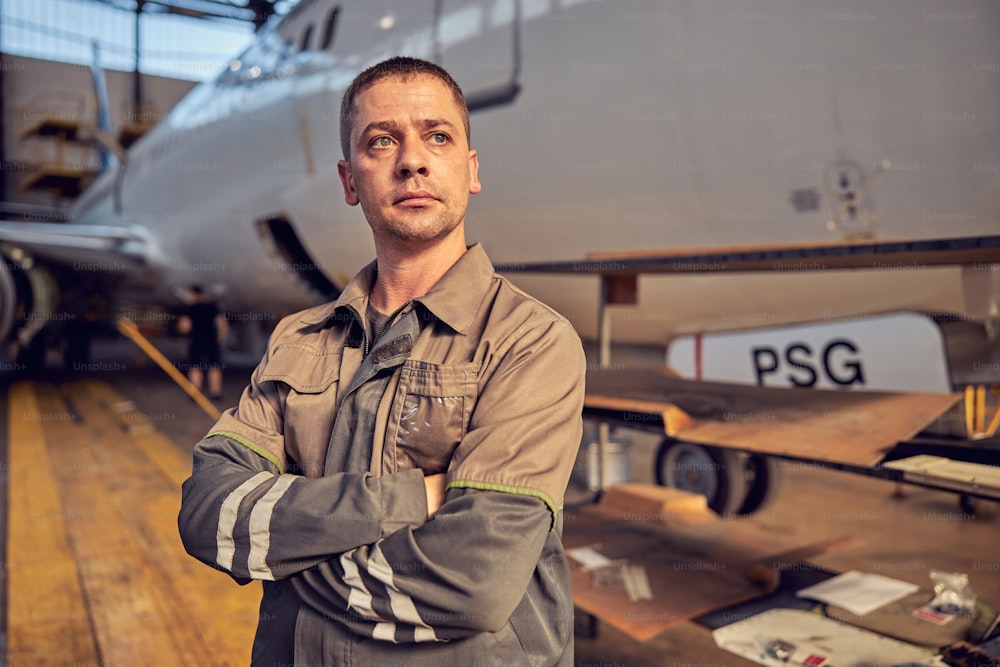 制服を着たハンサムで自信のある航空エンジニアの接写は、大きな民間航空機の前に立っている間、腕を組んでいます