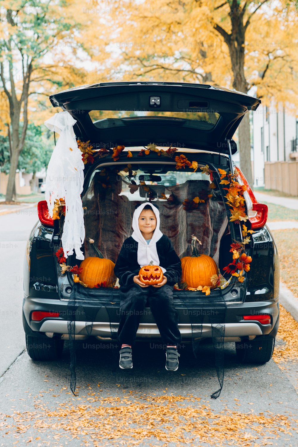 トリックかトランクか。車のトランクでハロウィンを祝う男の子。伝統的な10月の休日を屋外で祝う赤い彫刻のカボチャを持つ子供。ソーシャルディスタンスと安全な代替祝賀会。