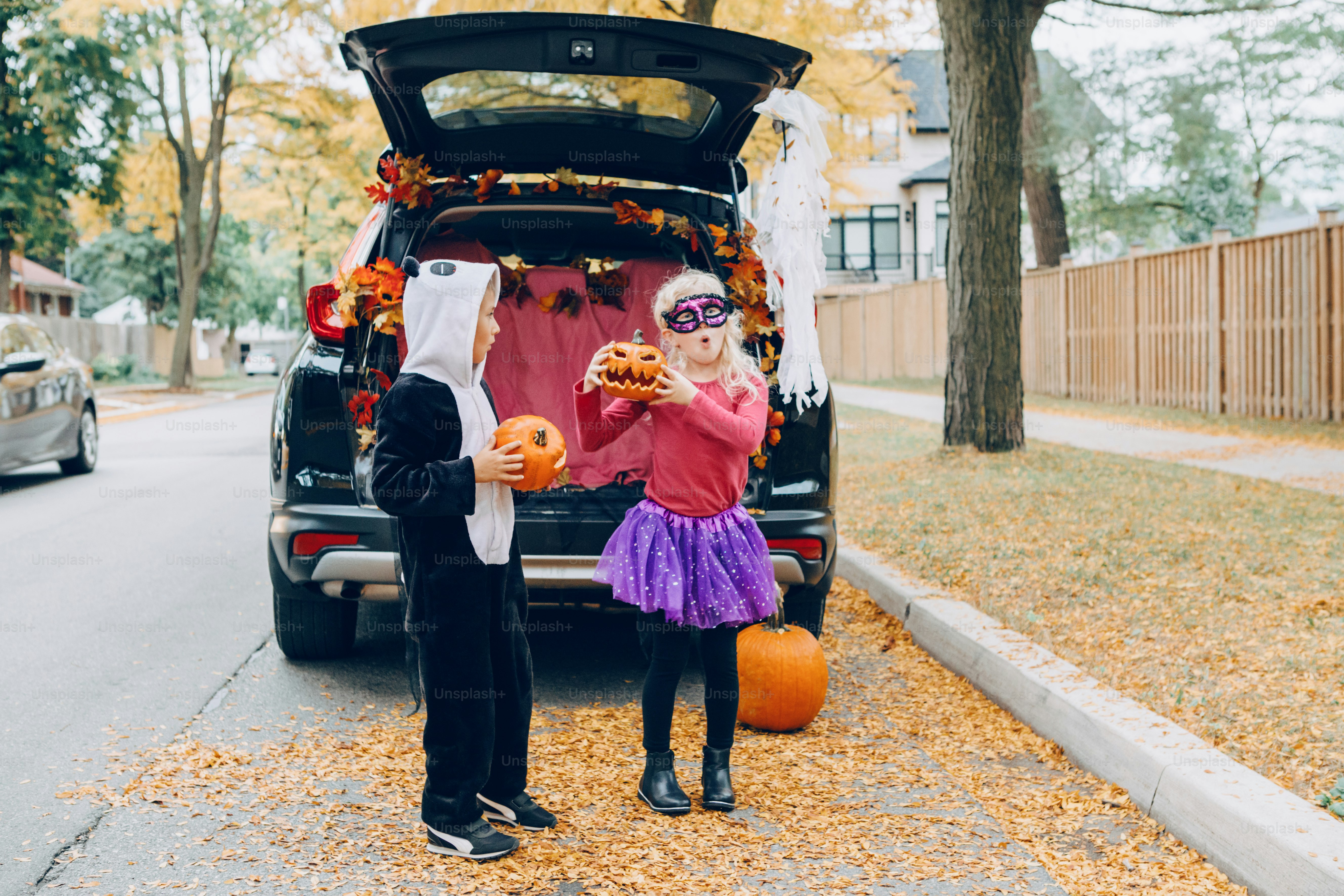Trucco o tronco. Bambini che festeggiano Halloween nel bagagliaio dell'auto. Ragazzo e ragazza con zucche rosse che celebrano la tradizionale festa di ottobre all'aperto. Distanziamento sociale e celebrazione alternativa sicura.