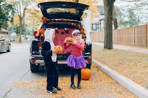 Trick or trunk. Children celebrating Halloween in trunk of car. Boy and girl with red pumpkins celebrating traditional October holiday outdoor. Social distance and safe alternative celebration.