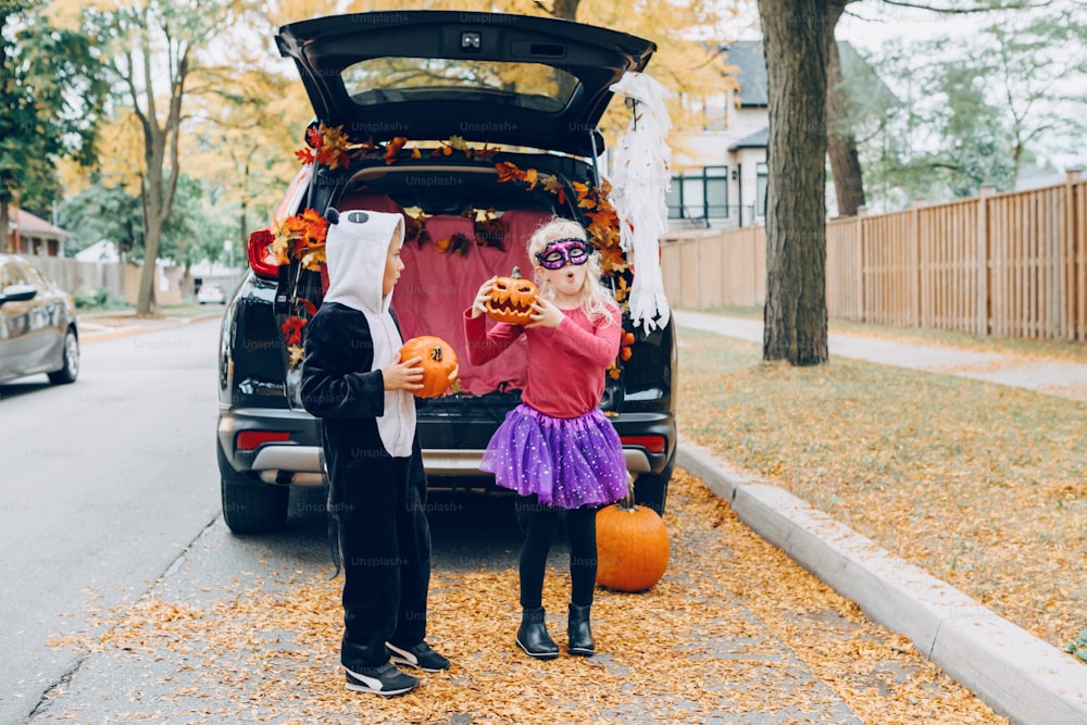 トリックかトランクか。車のトランクの中でハロウィンを祝う子供たち。伝統的な10月の休日を屋外で祝う赤いカボチャを持つ男の子と女の子。ソーシャルディスタンスと安全な代替祝賀会。