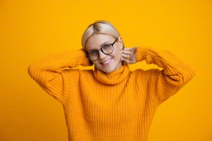 안경과 주황색 따뜻한 스웨터를 입은 금발 여성의 달콤한 흑백 초상화가 스튜디오 벽의 카메라에서 웃고 있다