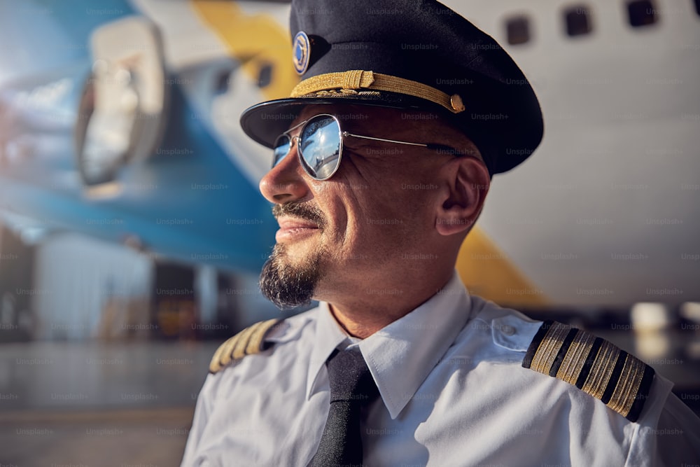 Retrato da vista lateral do belo piloto barbudo usando óculos escuros e uniforme de negócios olhando para o céu enquanto está perto da aeronave