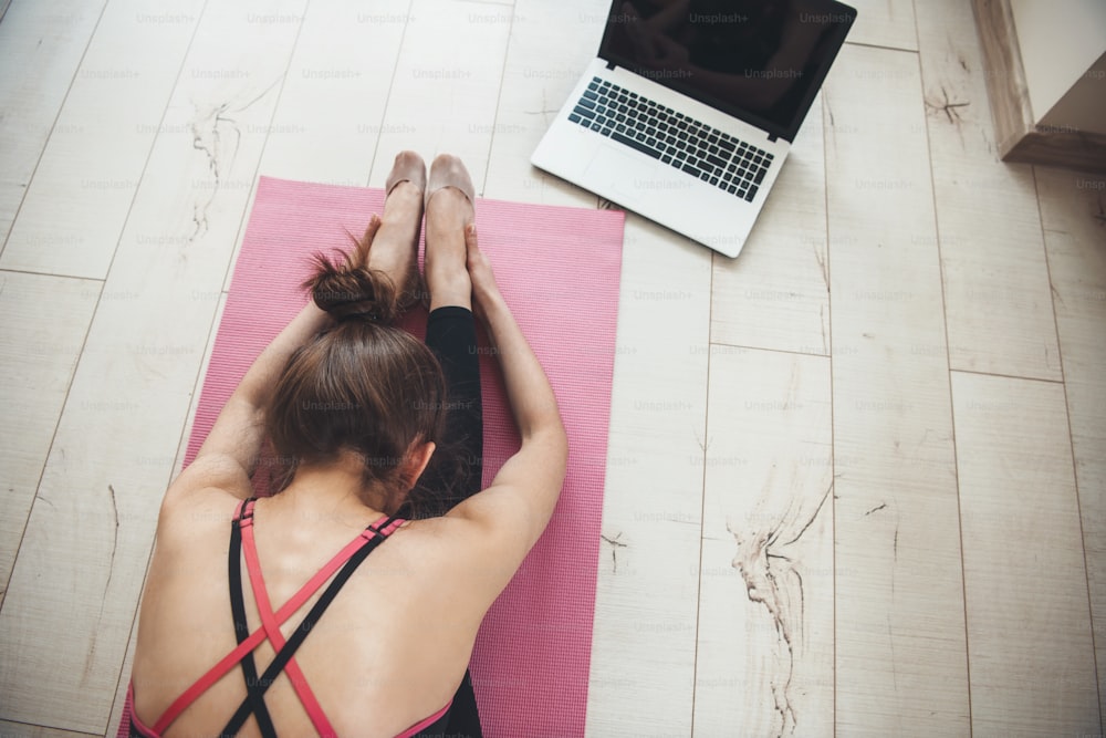 Upper view photo of a caucasian woman in sportswear doing yoga at home on the floor using a laptop