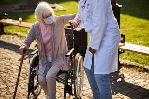 Foto recortada de una anciana que se levanta mientras se apoya en un bastón y sostiene la mano de un médico