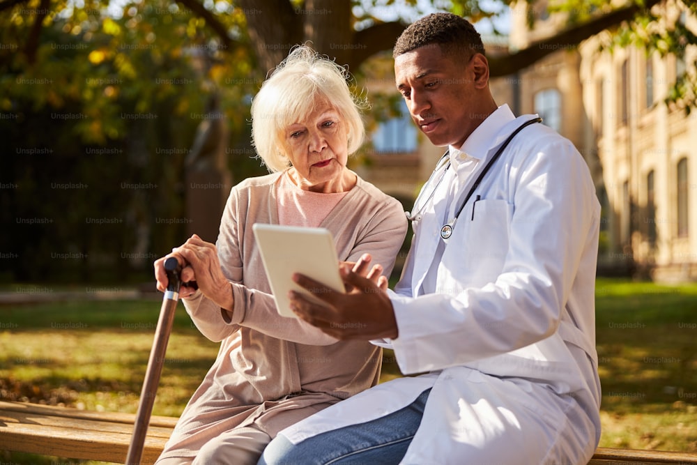 Youthful medical practitioner displaying useful information on his pad to a pensioner while sitting on a bench