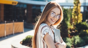 Freckled caucasian woman with red hair and eyeglasses embracing a laptop and posing at camera with a phone