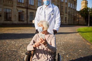 Aged woman with disability holding a smartphone and looking away while unrecognized doctor pushing her wheelchair