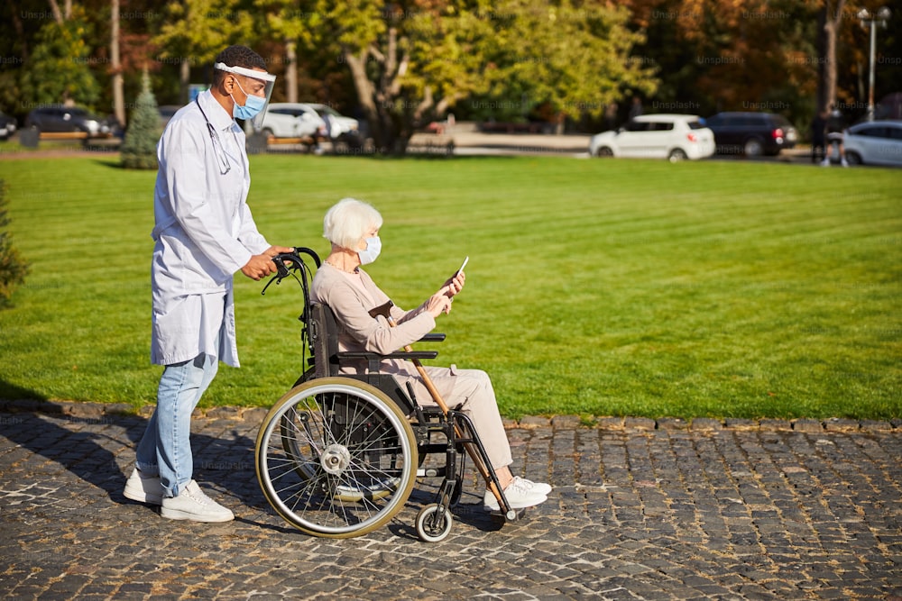 Elderly lady sitting with a smartphone in hands while a medical worker in protective shield pushing her wheelchair