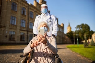 Arzt in Schutzschild und weißem Kittel schiebt den Rollstuhl einer älteren Frau mit modernem Smartphone