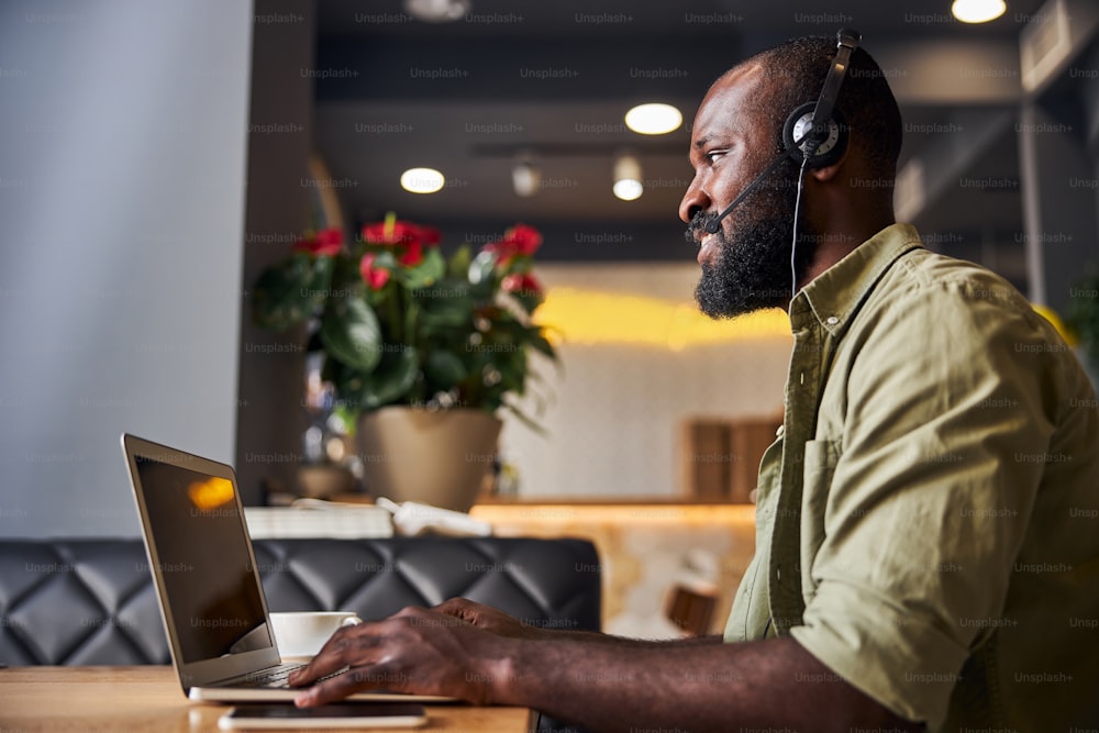 Smiling bearded gentleman wearing headset while sitting at the table with modern laptop