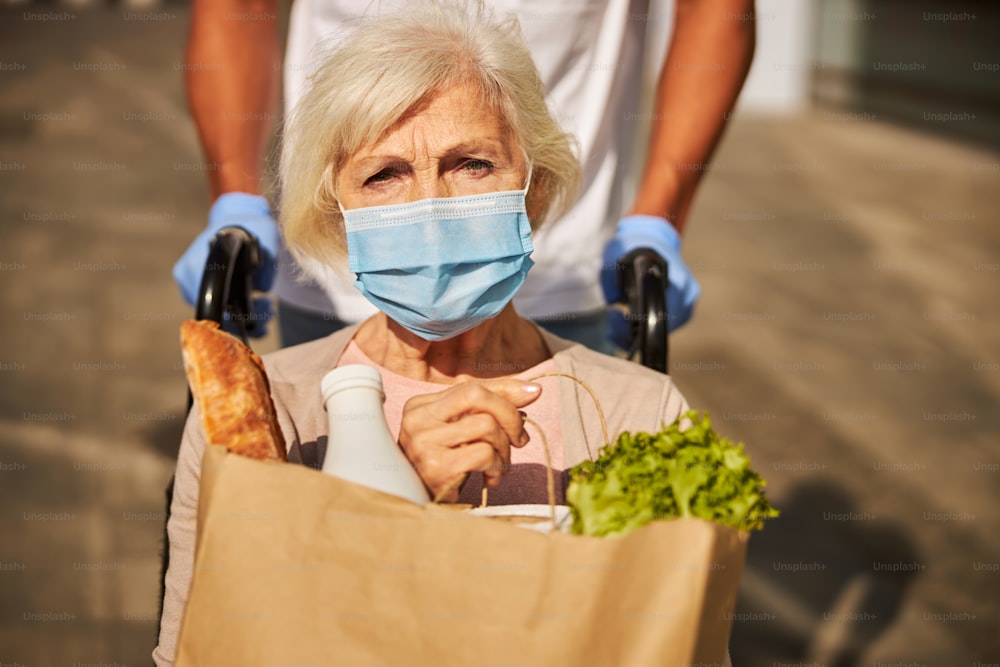 Foto de primer plano de una persona anciana con problemas con una máscara facial que guarda una bolsa marrón con baguette, leche y lechuga