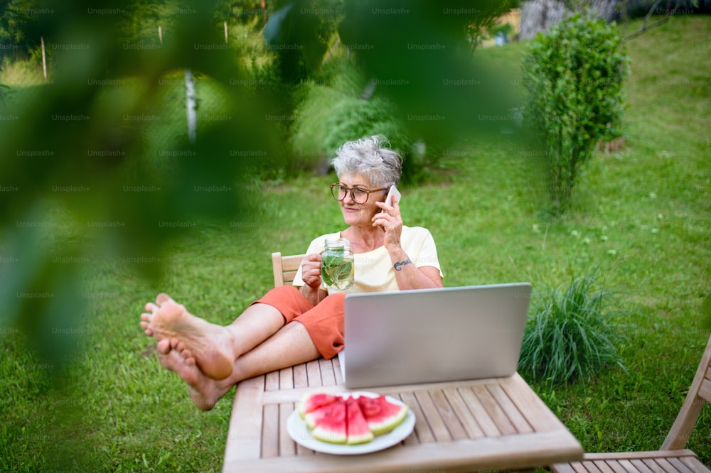 Mulher sênior feliz com laptop, smartphone e pés na mesa trabalhando ao ar livre no jardim, conceito de home office.