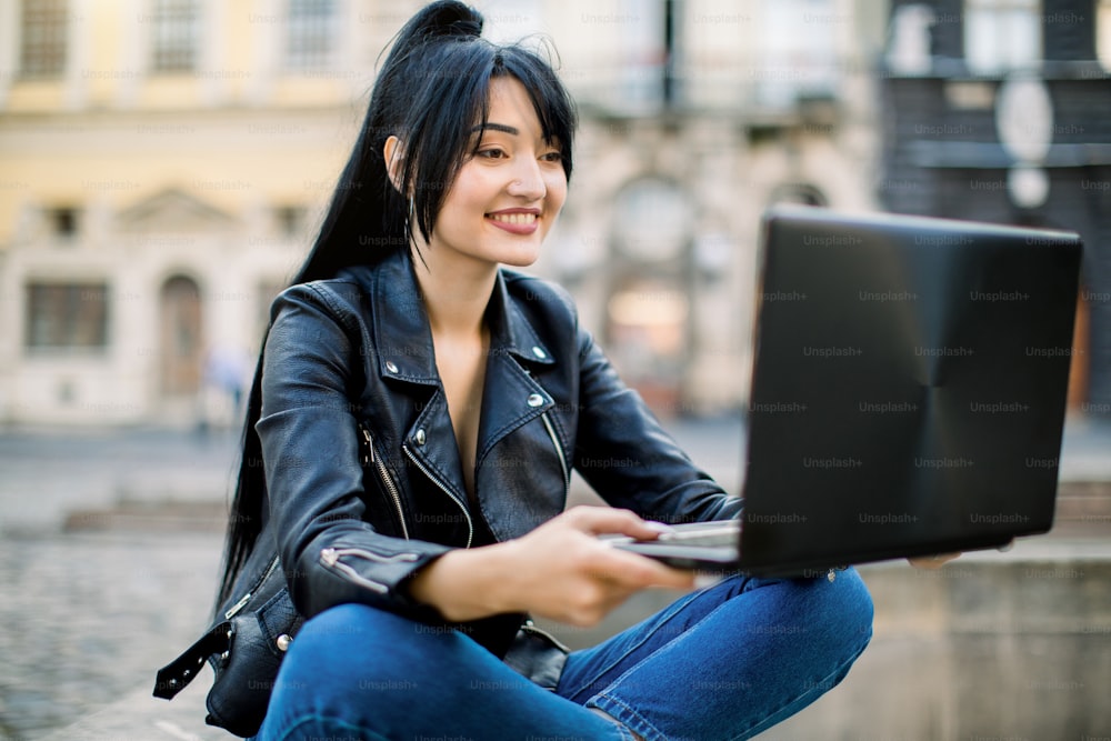 Image of casual pretty Asian brunette businesswoman sitting outdoor on vintage stone city fountain, while using her laptop and making video call. Business people, freelancers at work.