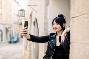 Foto urbana ao ar livre de uma bela mulher asiática sorridente jovem sexy em jaqueta de couro preta, fazendo autorretrato no smartphone, posando na rua da cidade velha. Selfie, pessoas, conceito de estilo de vida