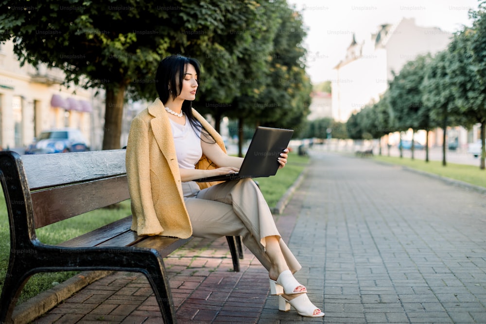 Pretty concentrated Asian businesswoman, wearing yellow blazer, sitting under trees on bench at city park and working on laptop. Bright morning sunlight on the background.