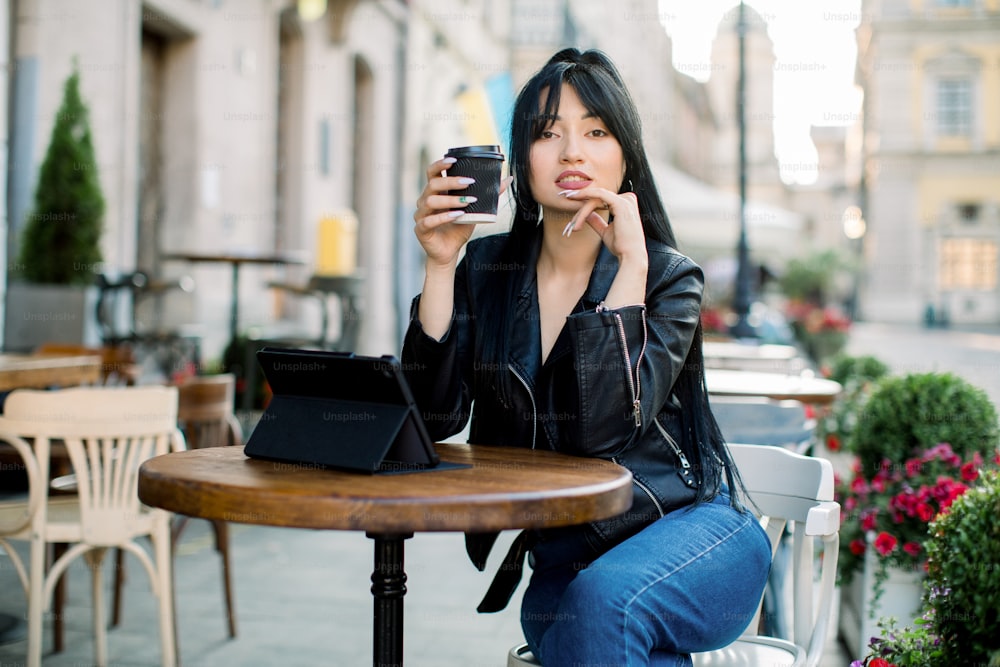 Concepto de estilo de vida freelance. Mujer asiática joven y trabajadora bonita con una tableta, bebiendo café en una cafetería al aire libre de la ciudad, trabajando en línea, leyendo noticias y disfrutando de la pausa para el café.