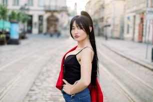 Young pretty mixed raced brunette girl in fashionable clothes, walking on the street of old city center, on sunny day weather, with ancient buildings on the background. City portrait of Asian woman.