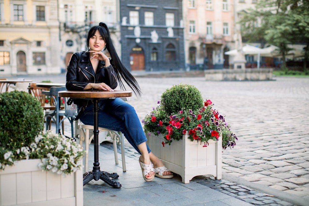Mujer morena asiática feliz en un café al aire libre, sentada a la mesa. Mujer joven con estilo en jeans y chaqueta de cuero, descansando al aire libre en el café en la vieja ciudad europea.